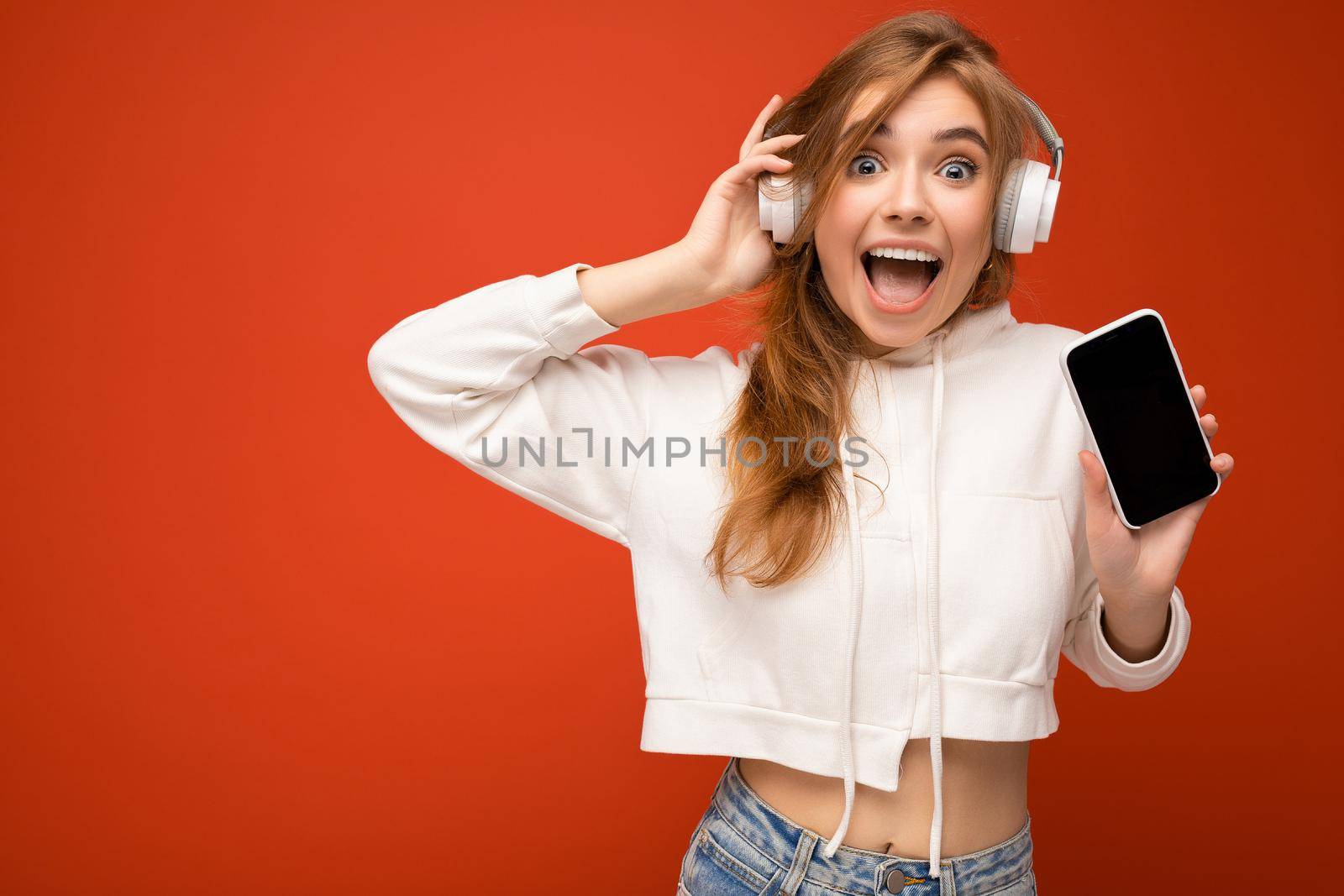 Beautiful positive amazed shocked young woman wearing stylish casual outfit isolated on background wall holding and showing mobile phone with empty display for mockup wearing white bluetooth headphones listening to music and having fun looking at camera.