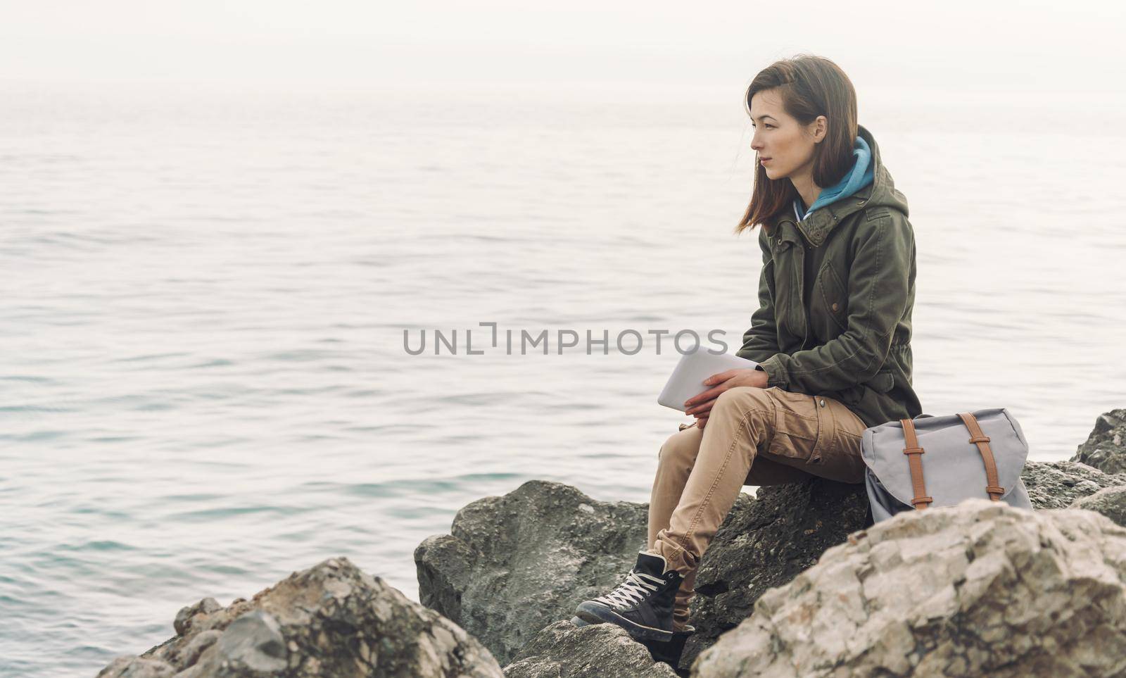 Traveler young woman with digital tablet and backpack sitting on stone coast