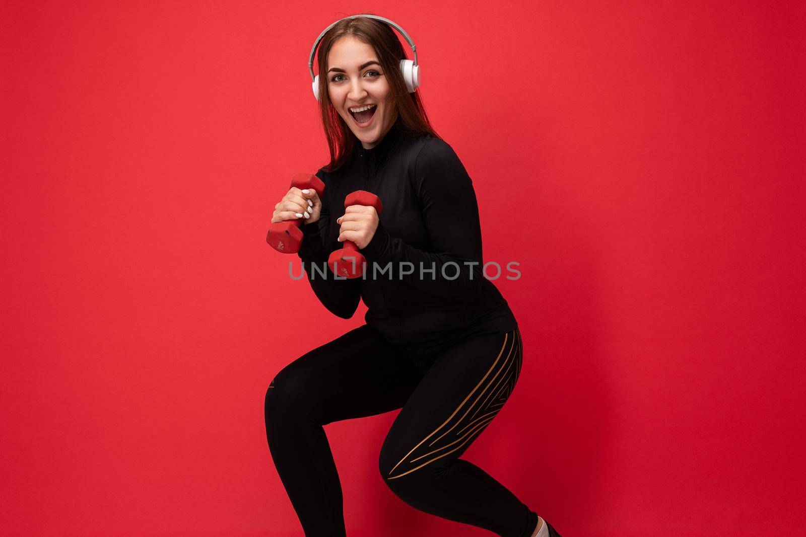 Side-profile photo of beautiful positive smiling young brunette female person wearing black sport clothes isolated on red background wall doing fitness using dumbbells wearing white bluetooth earphones listening to good music looking at camera.