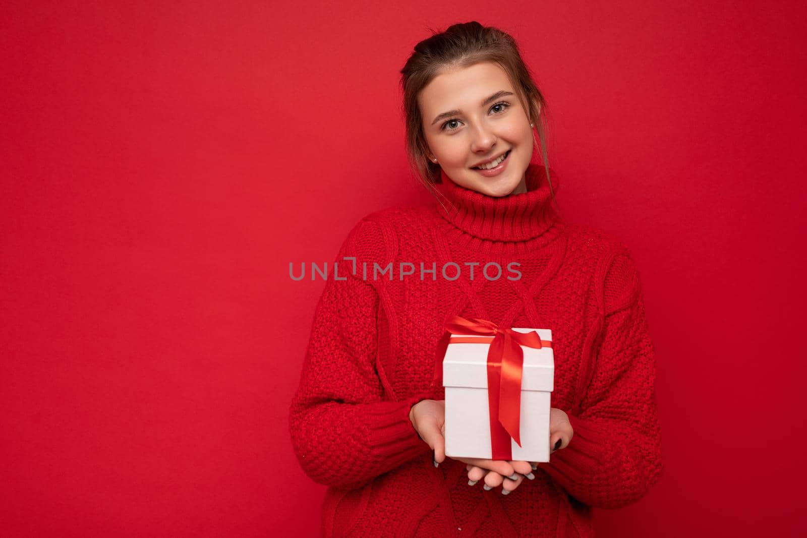 Beautiful happy young brunette woman isolated over colourful background wall wearing stylish casual clothes holding gift box and looking at camera. Free space