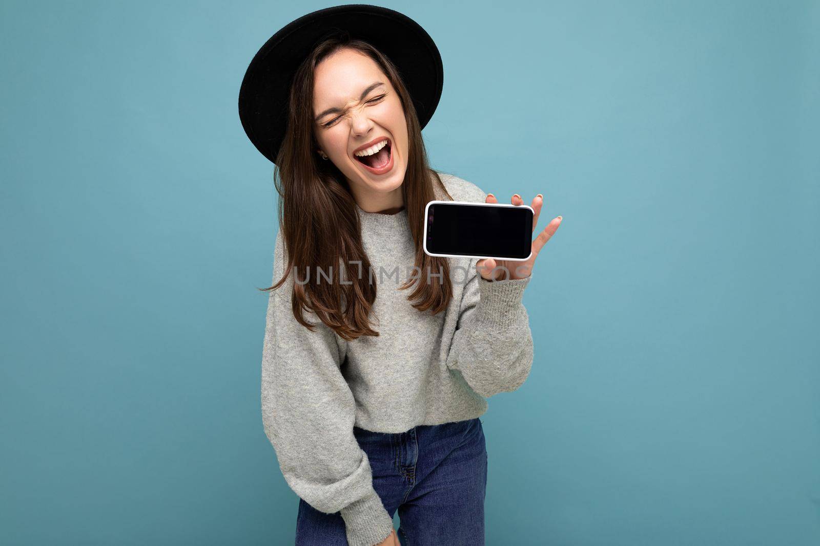 Portrait photo shot of beautiful young woman wearing black hat and grey sweater holding phone showing smartphone isolated on background with open mouth having joy.Mock up, cutout, copy space