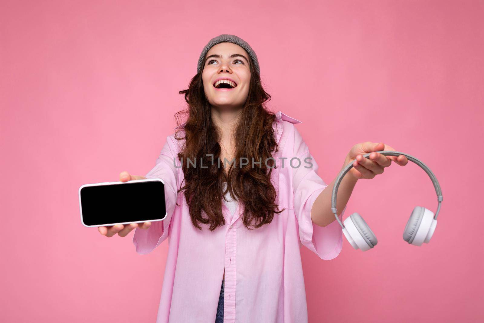 Photo of beautiful positive smiling young woman wearing stylish casual outfit isolated on colorful background wall holding white bluetooth wireless earphones and showing mobile phone with empty screen for mockup looking up by TRMK