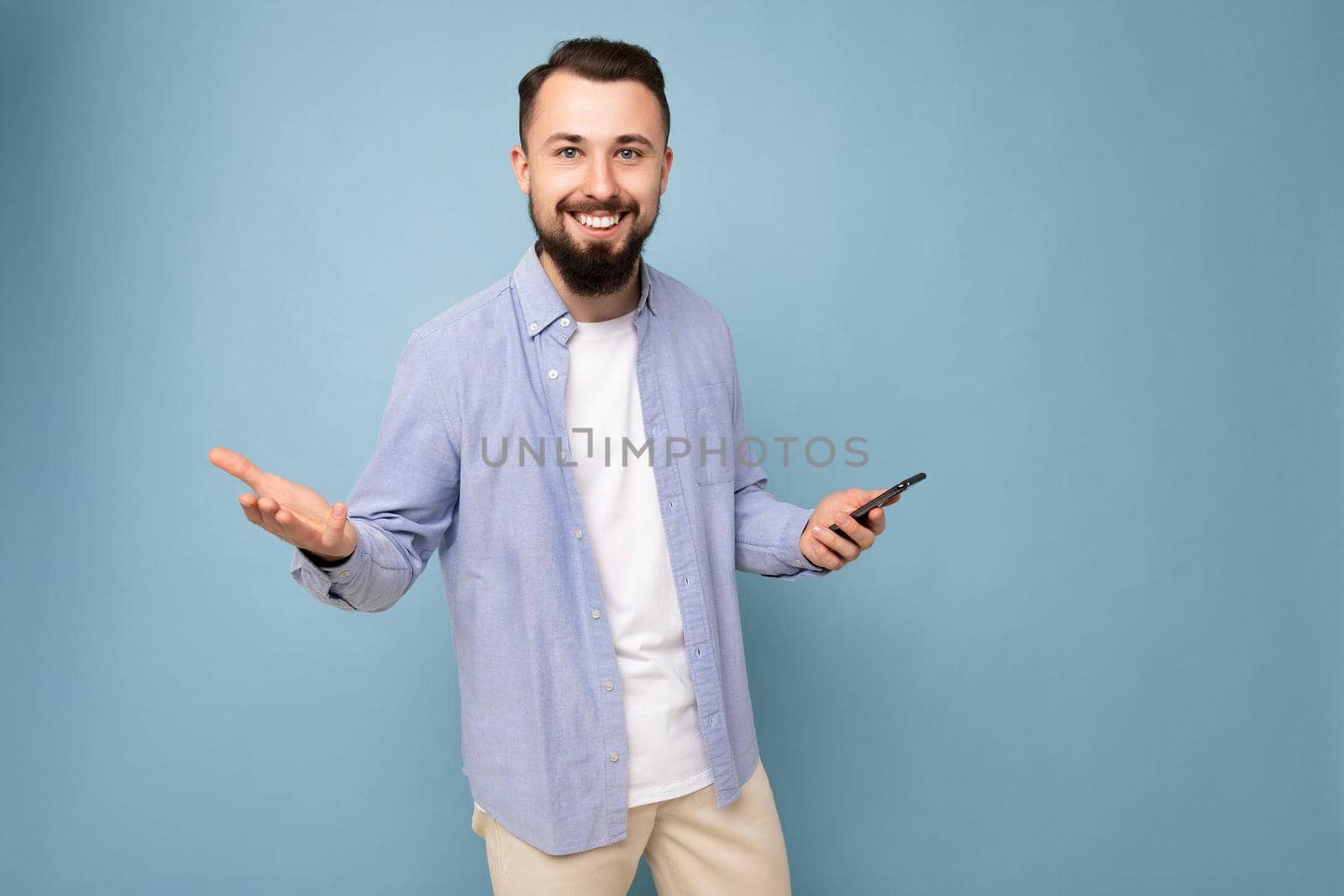 Happy smiling handsome young brunette unshaven man with beard wearing stylish white t-shirt and blue shirt isolated over blue background with empty space holding in hand and using phone messaging sms looking at camera.