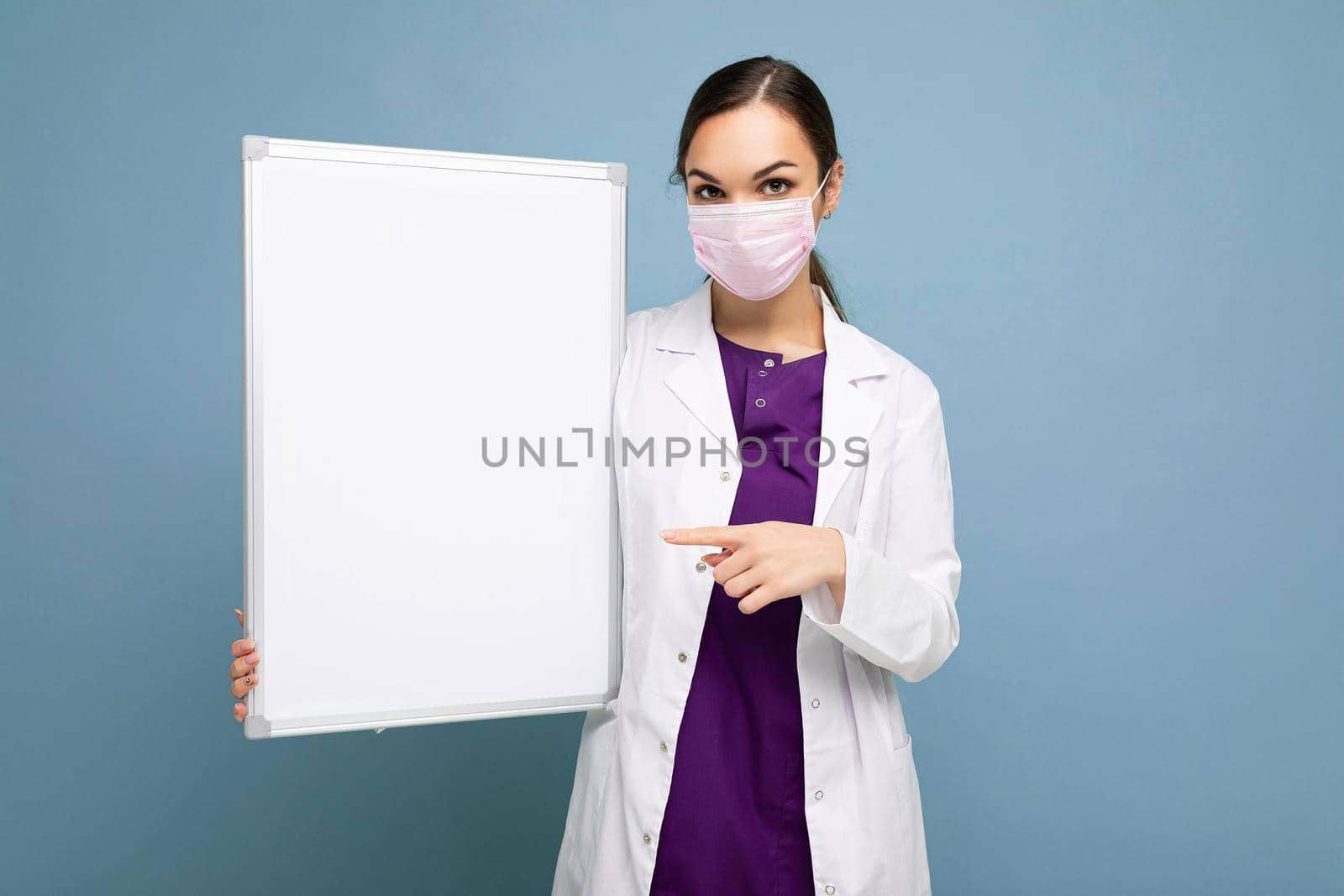 Photo of young attractive nurse in protective face mask and white medical coat holding an empty magnetic board isolated on blue background.
