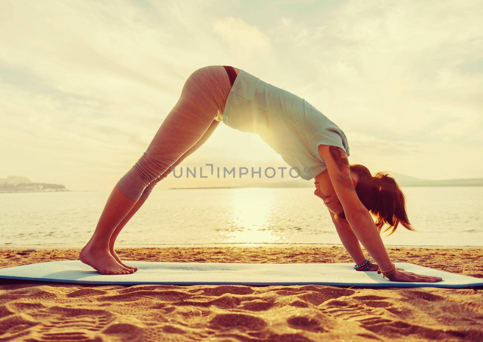 Young woman practicing yoga in downward facing dog pose on beach near the sea on sunset