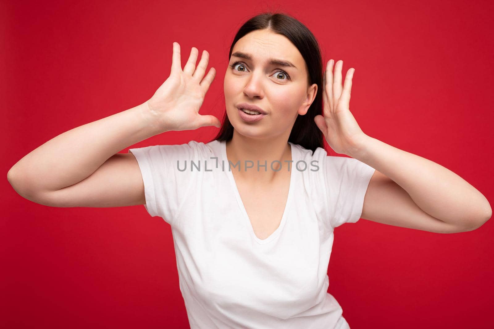 Portrait of emotional young beautiful brunet woman with sincere emotions wearing white t-shirt for mockup isolated over red background with empty space and covering ears.