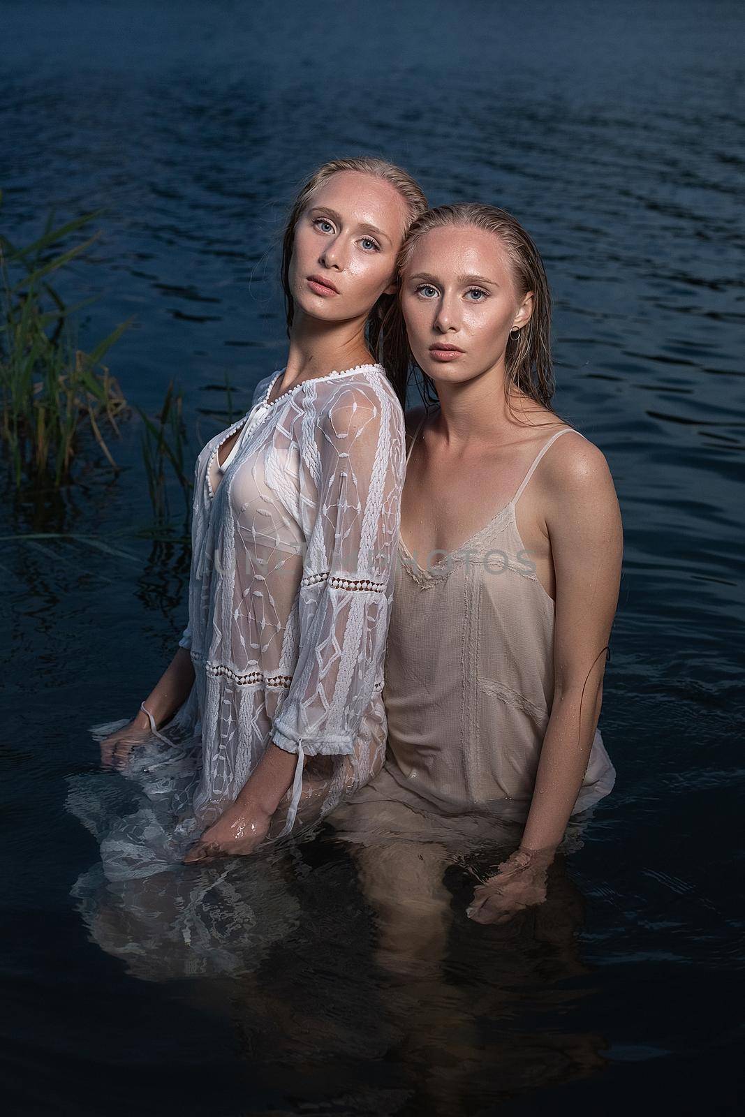 two young twin sisters with long blond hair posing in light dresses in water of lake at summer night, looking at camera. stylish fashion photoshoot with flashlight. outdoors evening photosession