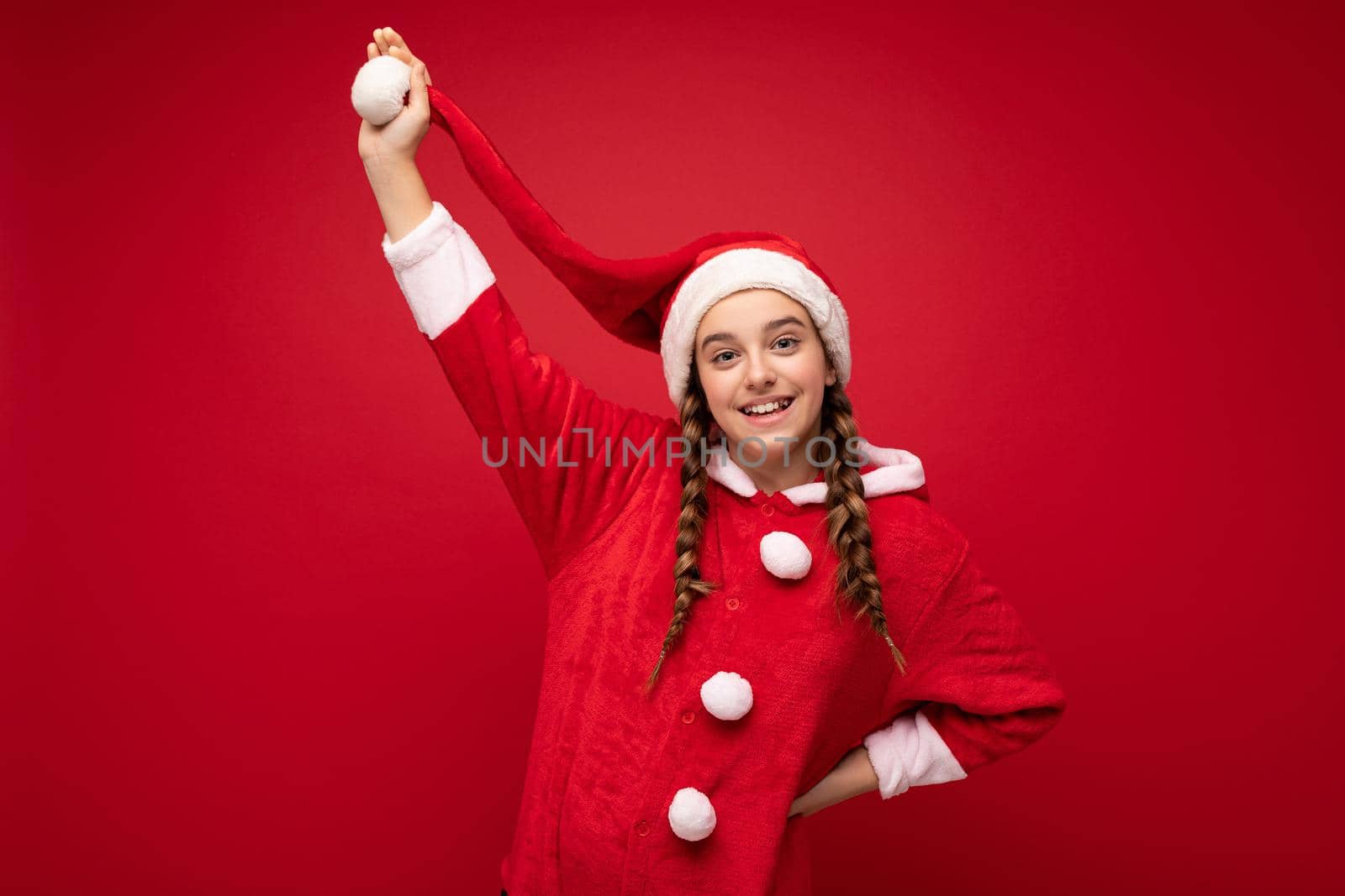 Photo shot of beautiful happy positive smiling brunette girl with pigtails wearing santa clause clothes isolated over red background wall looking at camera and having fun by TRMK