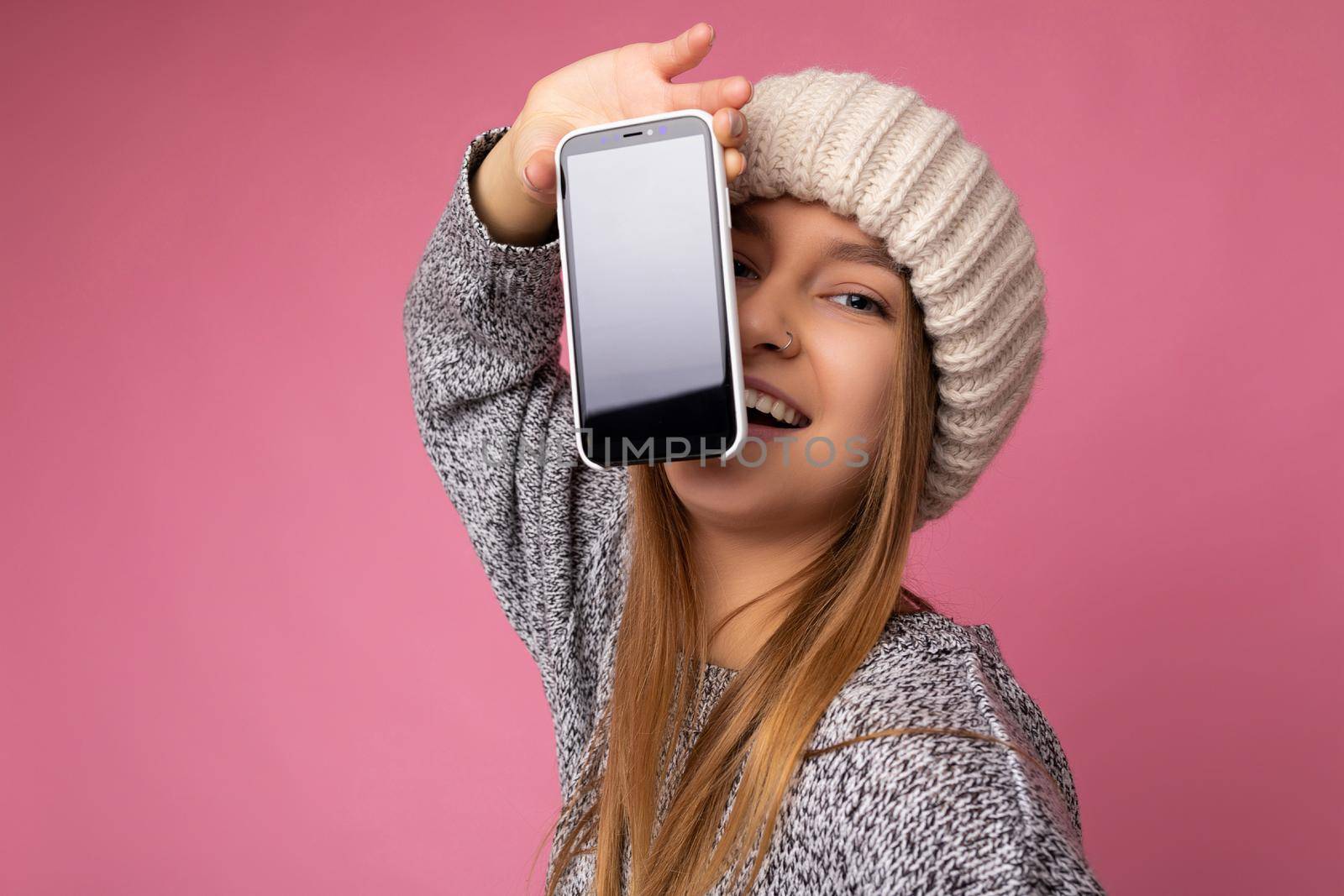photo of Beautiful young blonde woman wearing casual grey sweater and beige hat isolated over pink background holding in hand and showing mobile phone with empty display for mockup looking at camera.