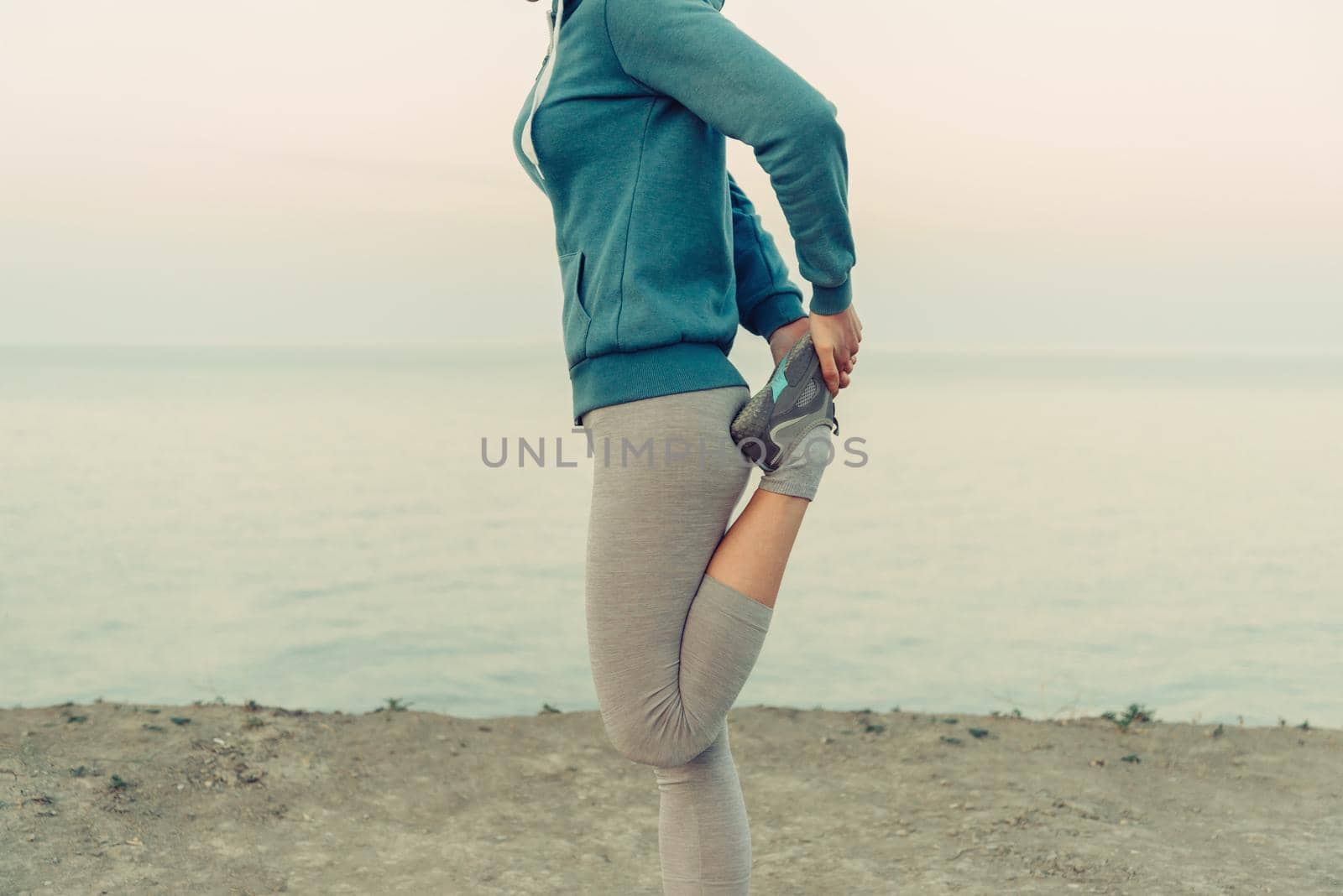 Young woman stretching her legs and preparing to run on coast in summer in the morning, face is not visible. Concept of sport and healthy lifestyle