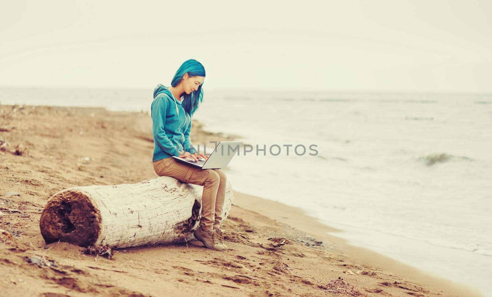 Freelancer young woman sitting on tree trunk and working on laptop on beach near the sea. Freelance concept