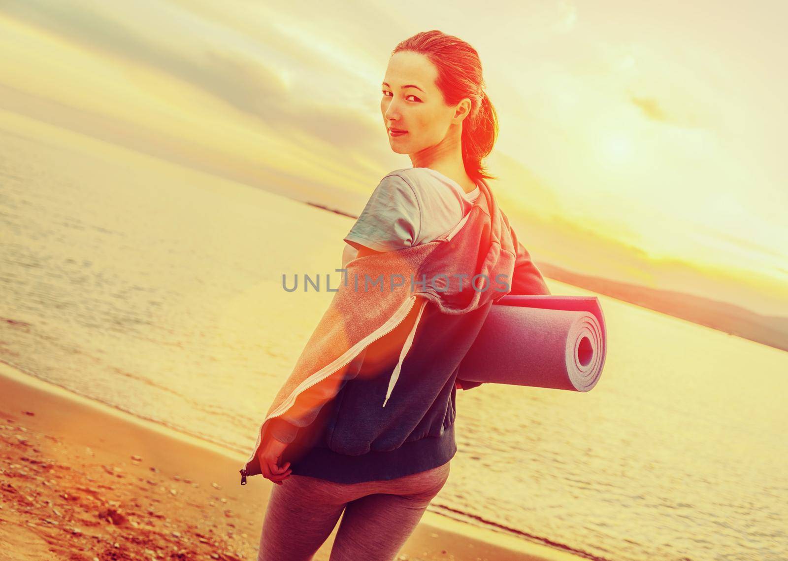Young beautiful woman with yoga mat standing on beach in summer at sunset and ready for workout. Image with sunlight effect