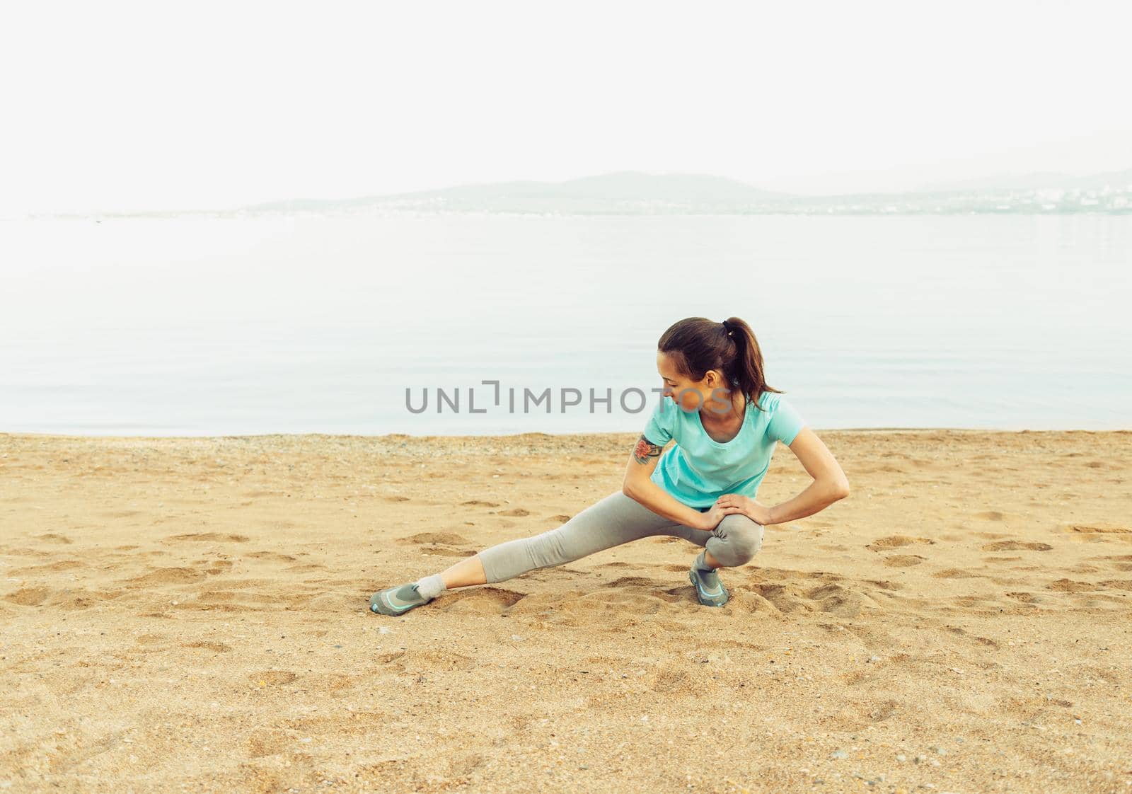 Young woman stretching her legs and preparing to run on beach near the sea in summer. Concept of healthy lifestyle