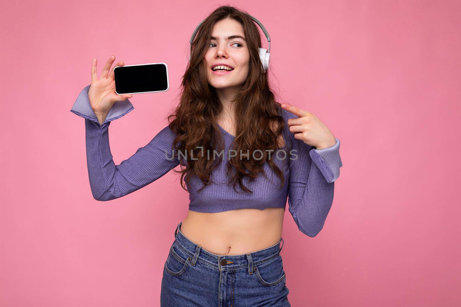 Attractive positive smiling young woman wearing stylish casual outfit isolated on colourful background wall holding and showing mobile phone with empty screen for cutout wearing white bluetooth headphones and having fun looking to the side.