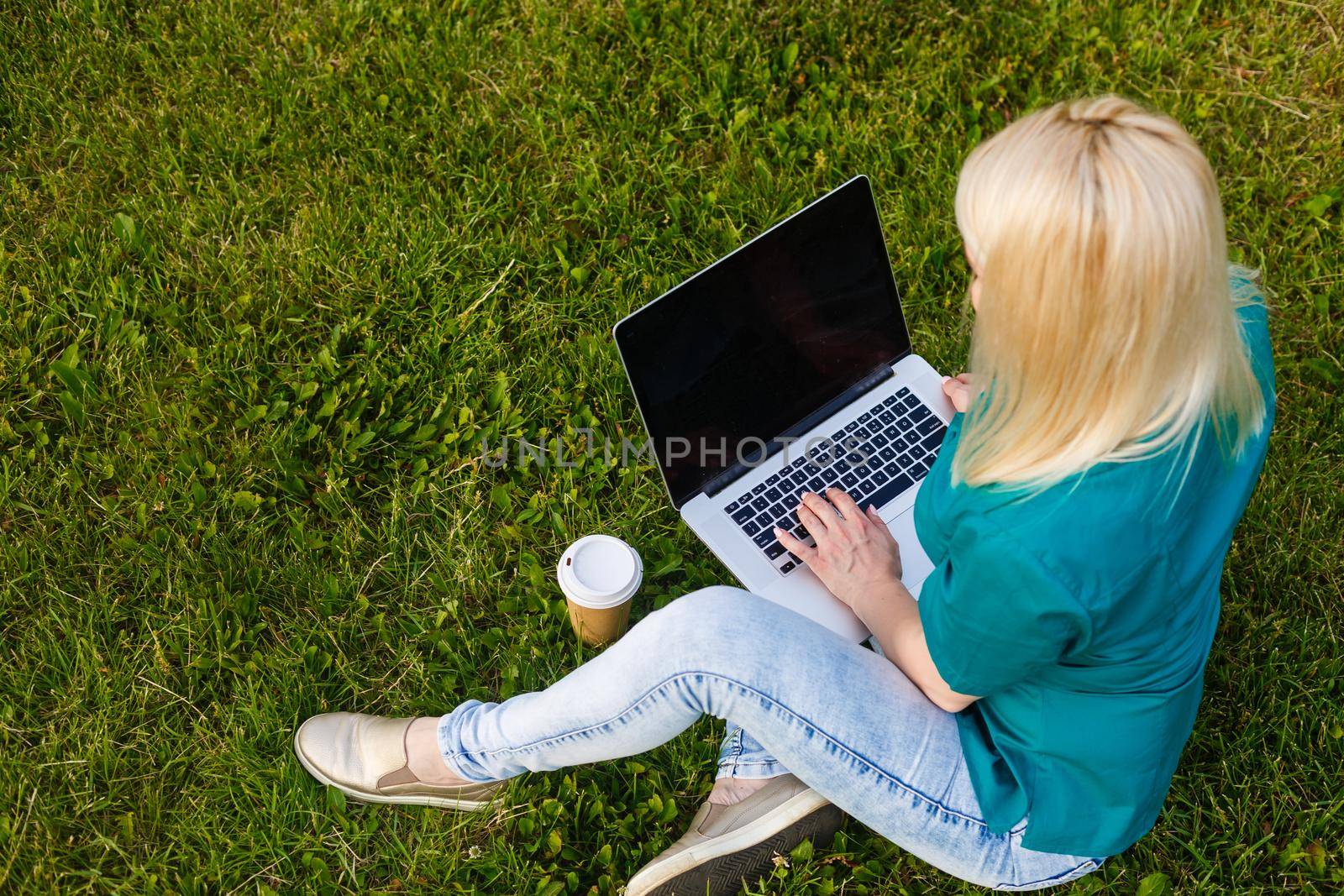 Top view of woman sitting in park on the green grass with laptop, hands on keyboard. Computer screen mockup. Student studying outdoors. Copy space for text