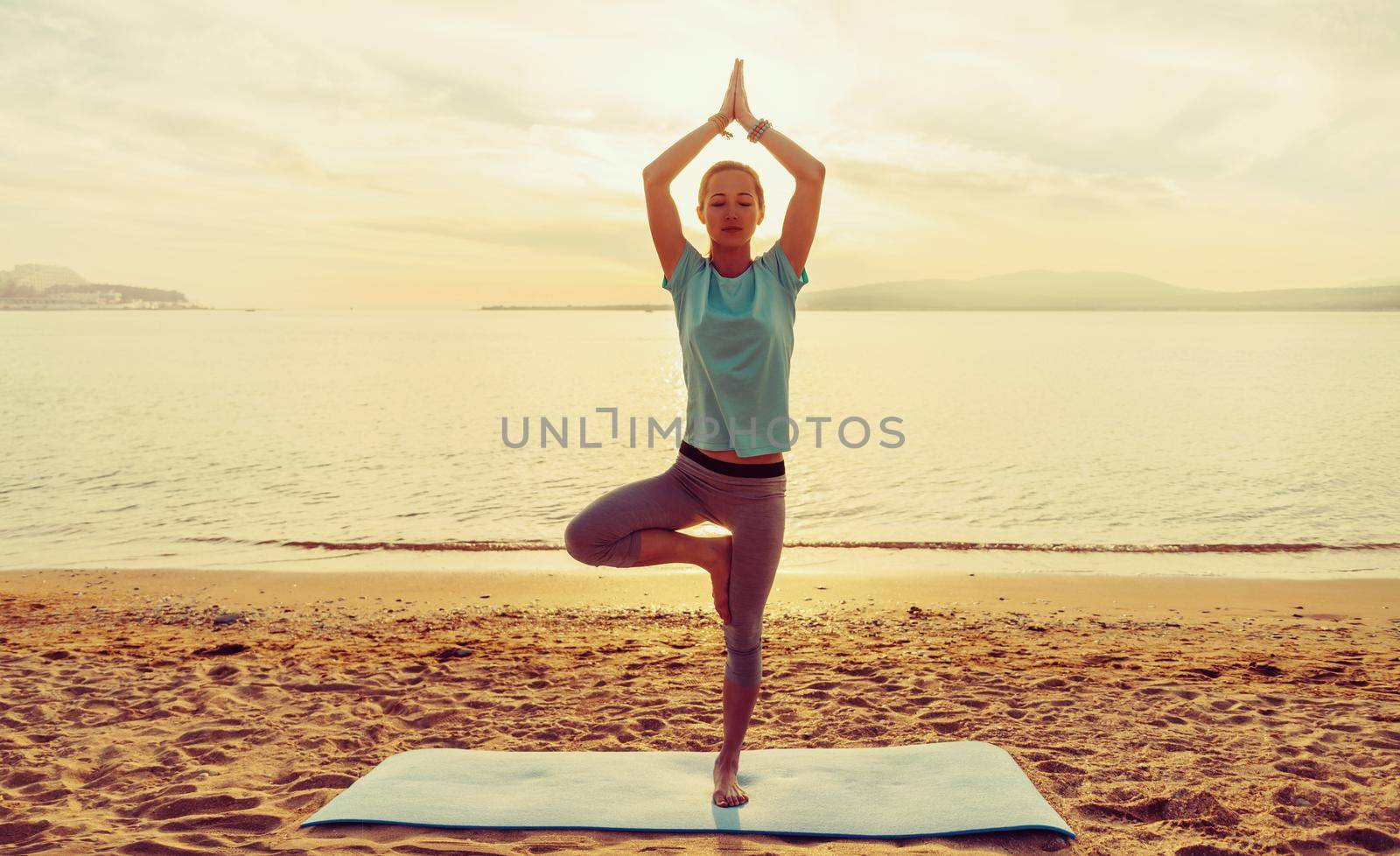 Young woman practicing yoga in pose of tree on beach near the sea at sunset in summer, front view