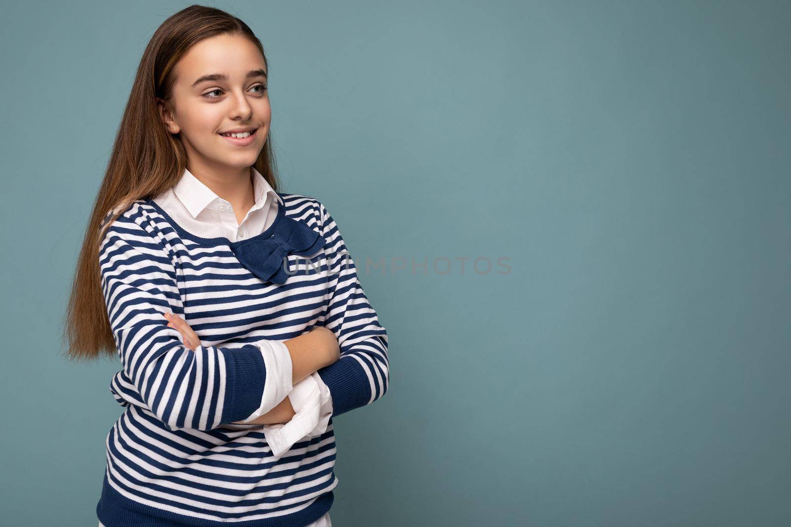 Shot of beautiful positive smiling brunette little girl wearing striped longsleeve standing isolated over blue background wall crossing hands and looking to the side by TRMK