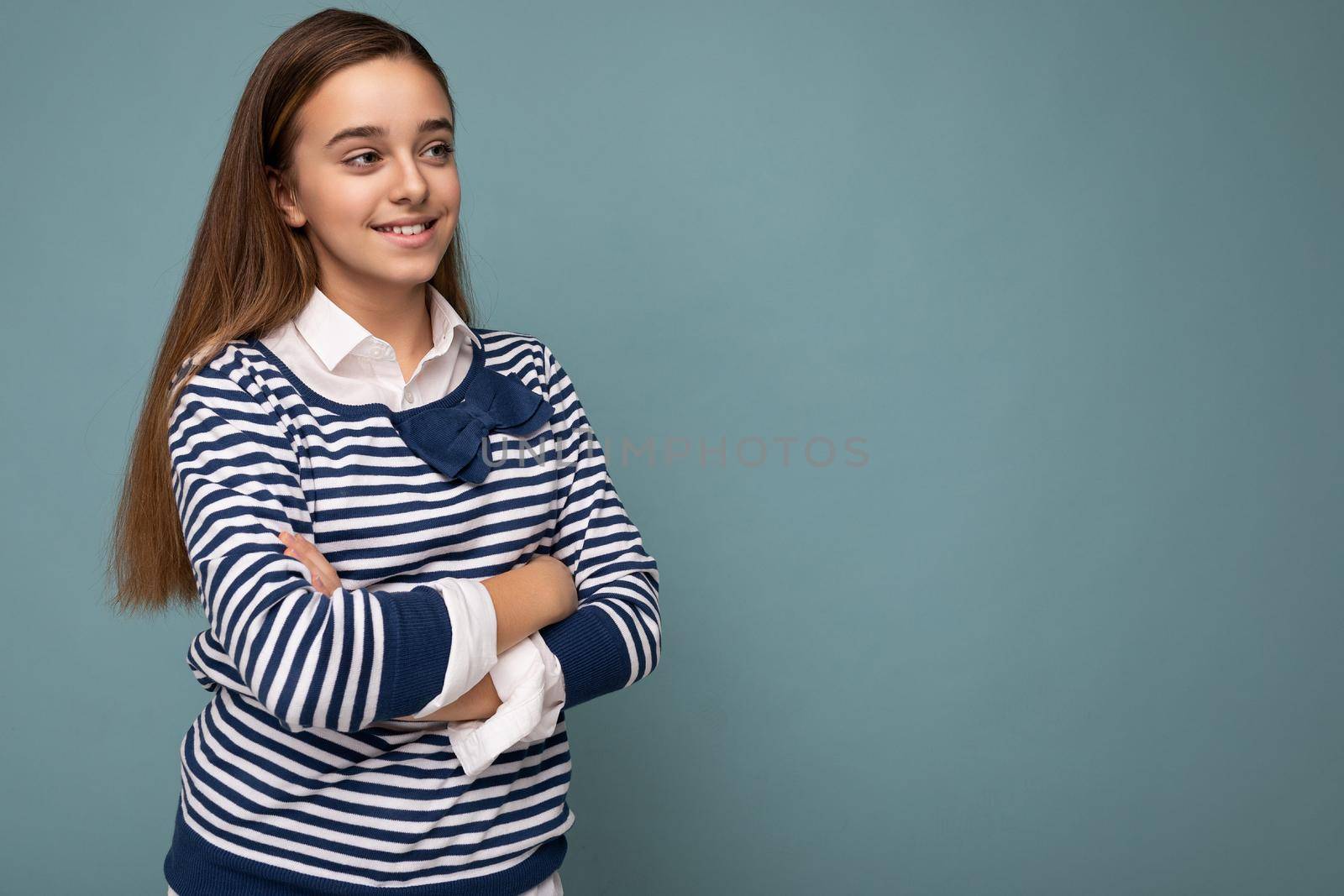 Shot of beautiful positive smiling brunette little girl wearing striped longsleeve standing isolated over blue background wall crossing hands and looking to the side. Copy space