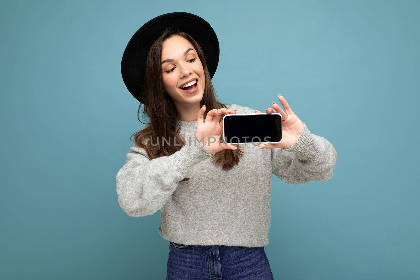 Attractive young smiling woman wearing black hat and grey sweater holding smartphone looking down isolated on background.Mock up, cutout