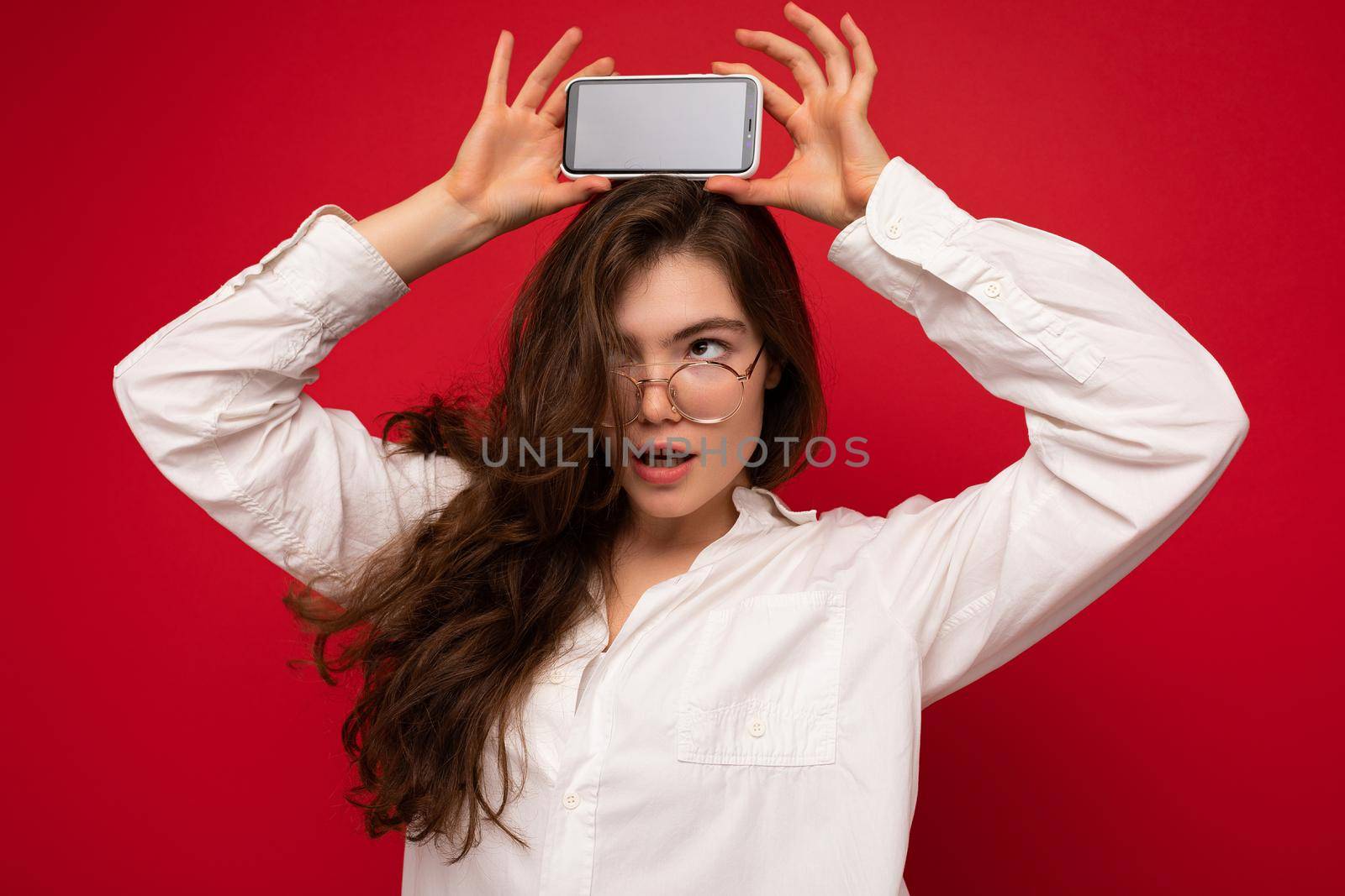photo of sexy beautiful young brunette woman wearing white shirt and optical glasses isolated over red background holding in hand and showing mobile phone with empty screen for cutout looking at gadjet screen.