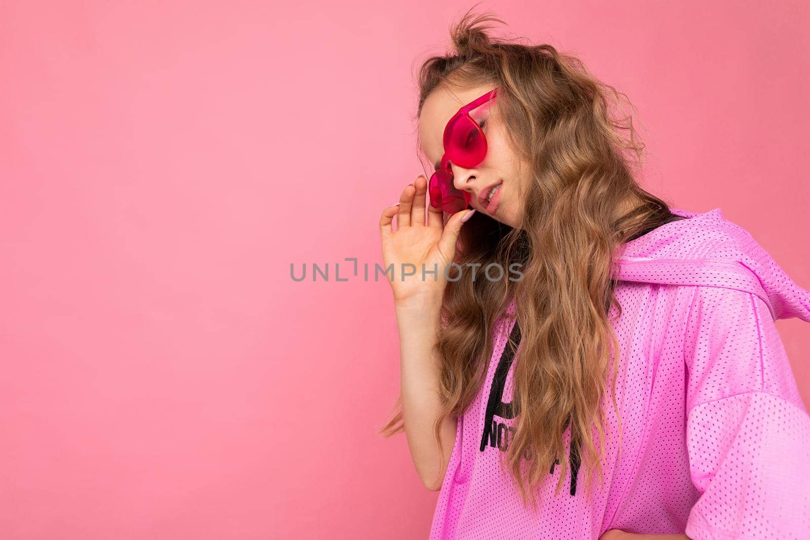 Side-profile photo shot of charming sorrowful young blonde curly woman isolated on pink background wall wearing casual pink t-shirt and stylish colourful sunglasses looking to the side by TRMK