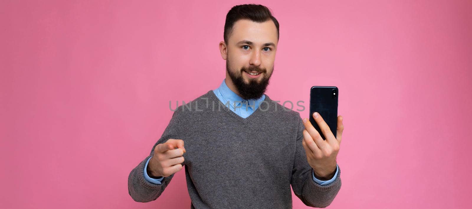 Panoramic photo of Handsome happy cool young brunette unshaven man with beardwearing stylish grey sweater and blue shirt standing isolated over pink background wall holding smartphone and using phone looking at camera pointing finger at you.