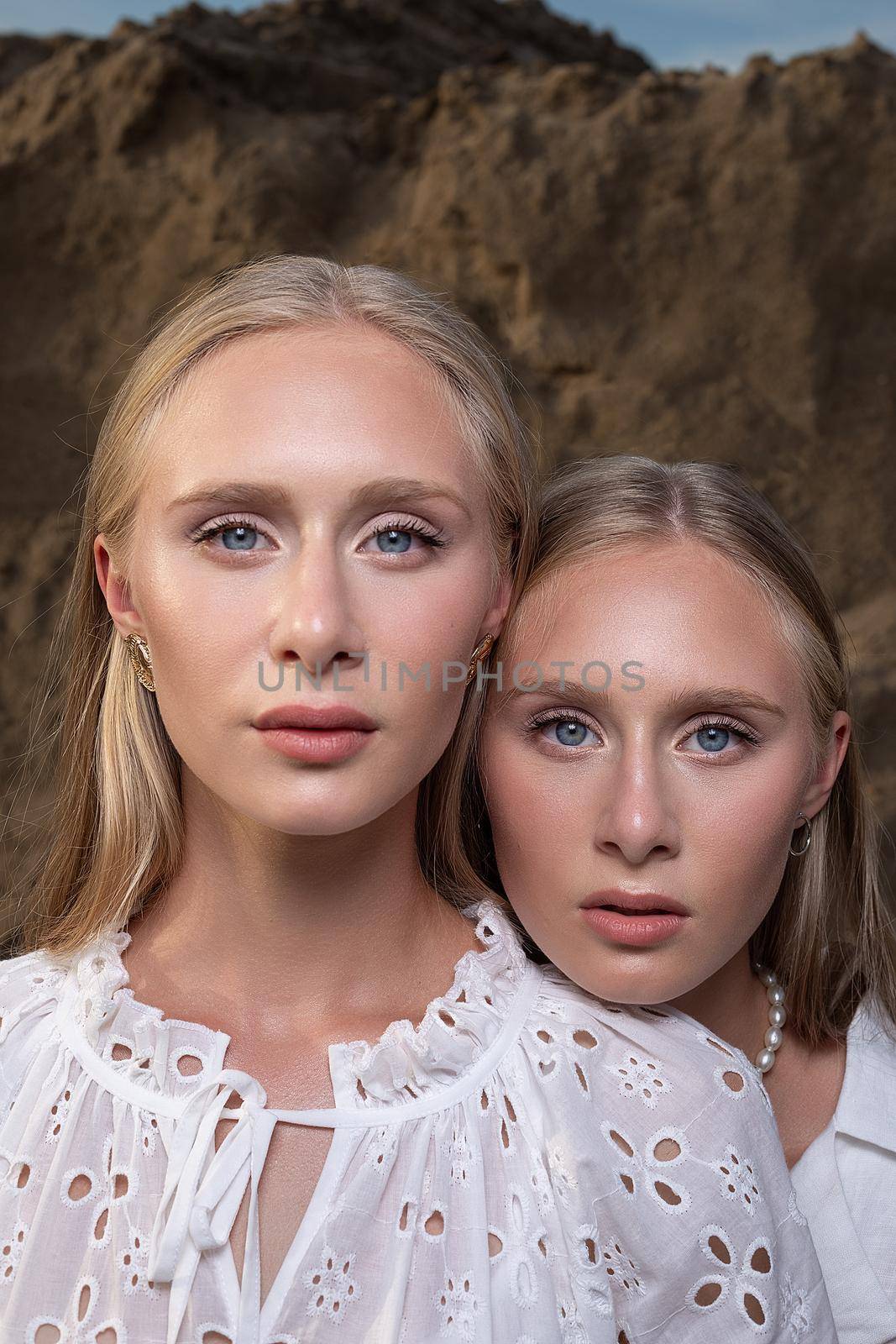 two young blond pretty twins posing at sand quarry in elegant white clothes by artemzatsepilin