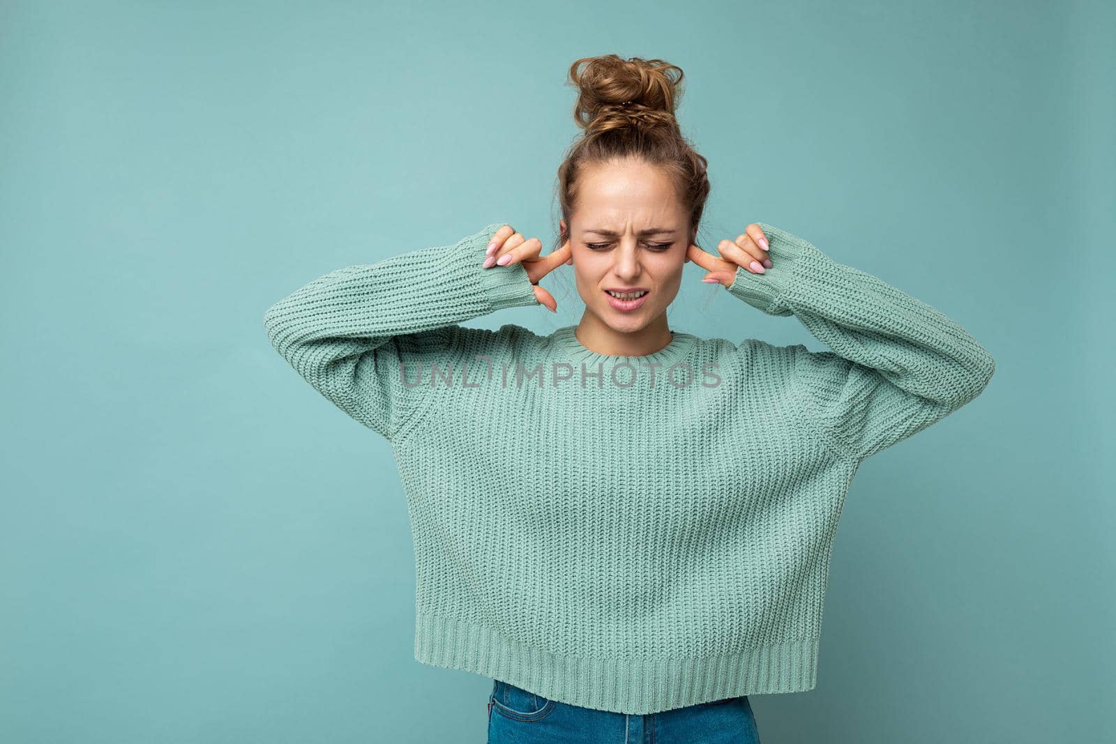 Portrait of young emotional nice winsome blond woman with sincere emotions wearing trendy blue sweater isolated over blue background with free space and covering ears by TRMK