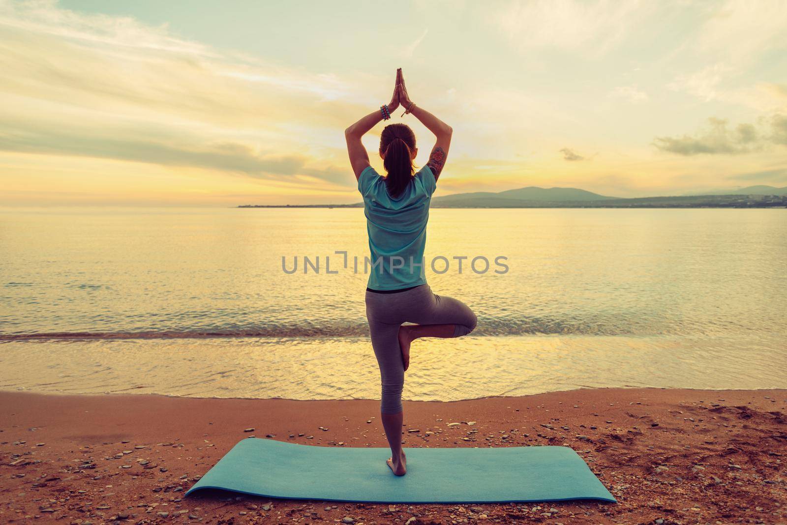 Young woman doing yoga exercise in pose of tree on beach at sunset in summer, rear view