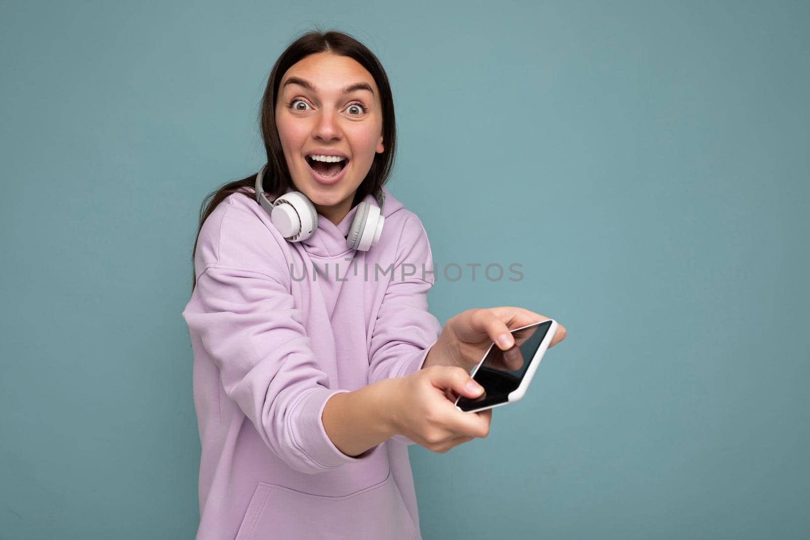 Beautiful positive smiling surprised young brunet female person wearing purple hoodie isolated on blue background holding and using smartphone wearing white wireless earphones looking at camera having fun by TRMK