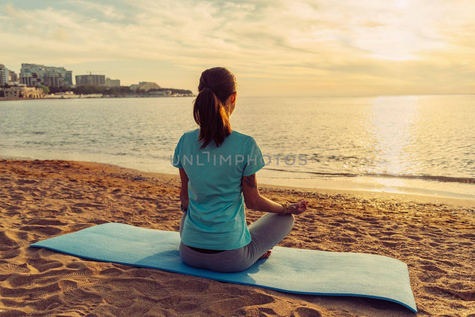 Young woman meditating in pose of lotus on sand beach near the sea at sunset in summer