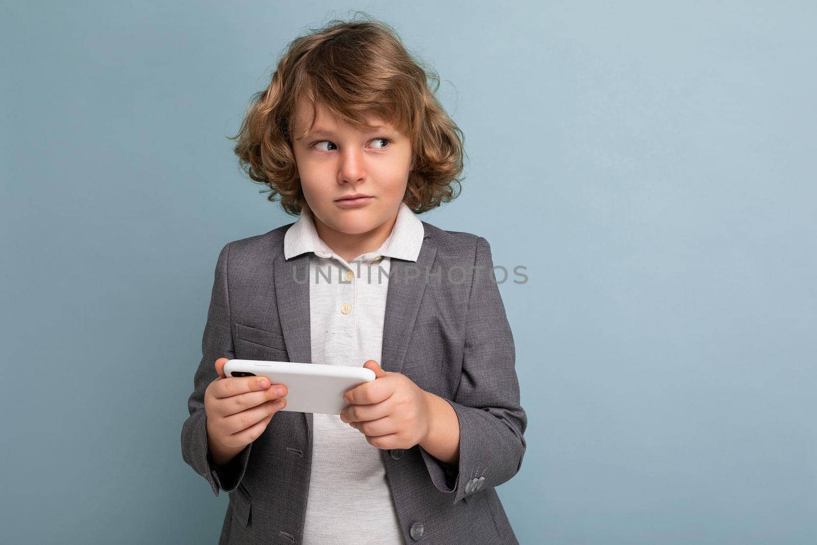 Photo shot of Handsome suspacted child boy with curly hair wearing grey suit holding and using phone isolated over blue background looking to the side playing games by TRMK