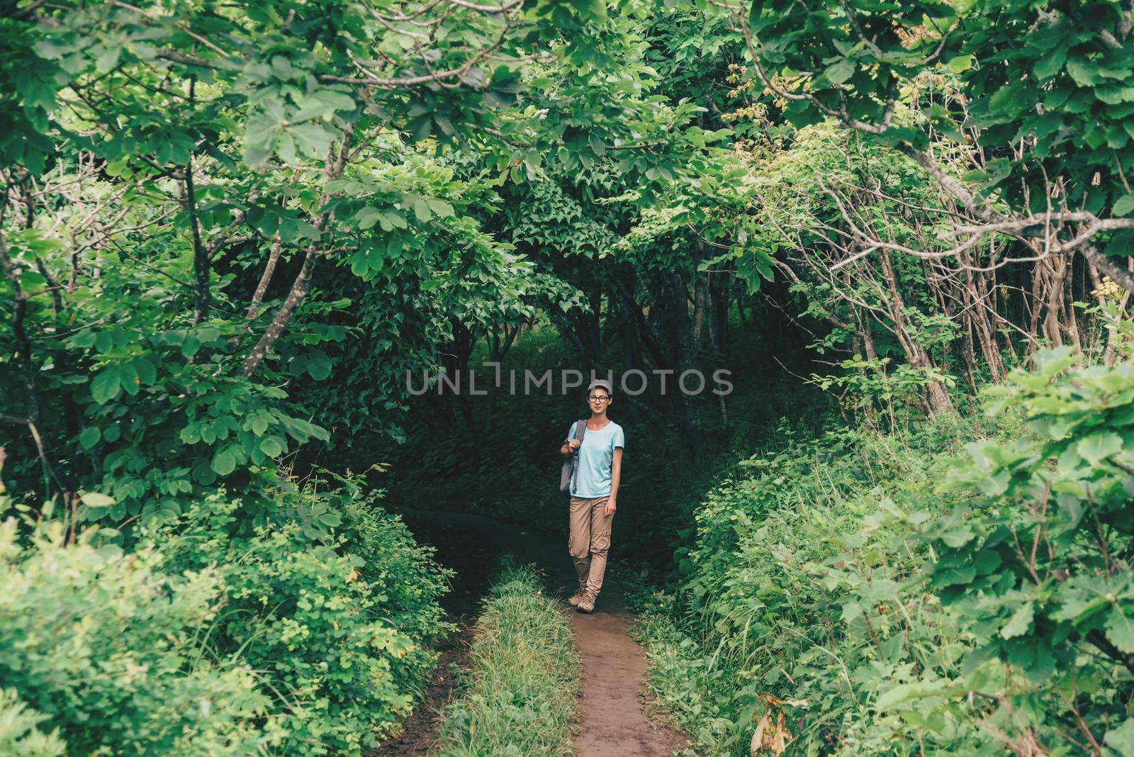 Traveler young woman walking in beautiful summer forest. Hiker girl goes out from the tunnel of green trees, girl looking at camera