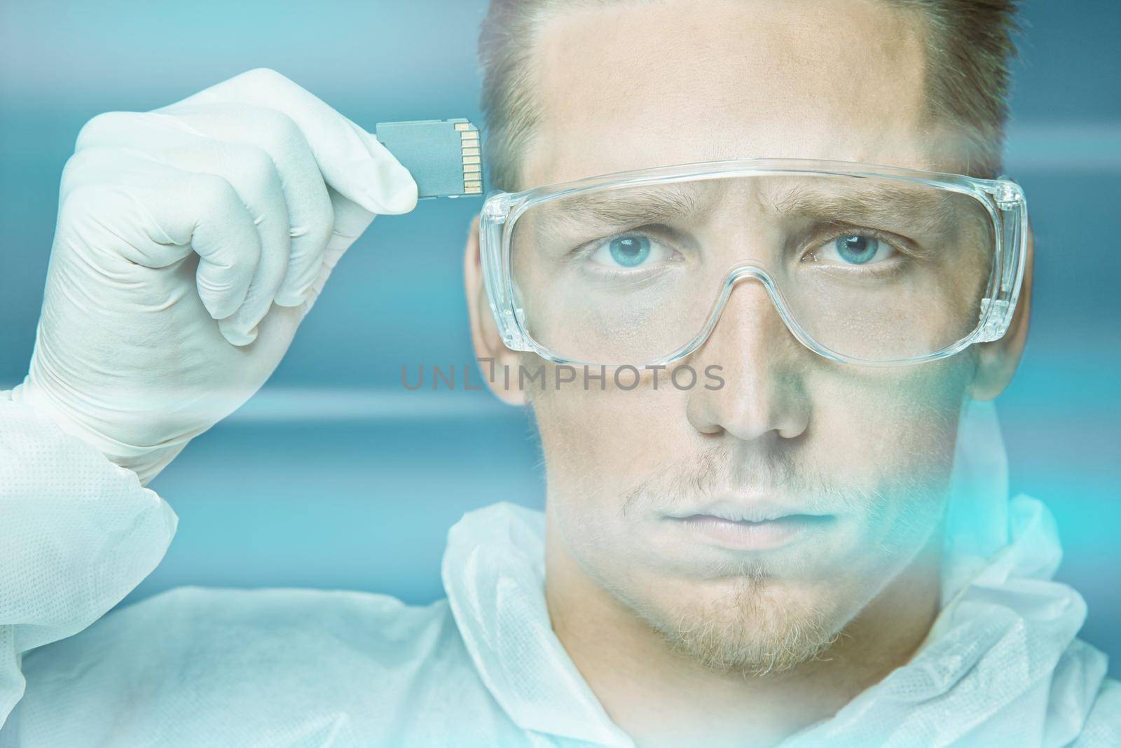 Serious young man in futuristic clothing holding a memory card near his head, cyber space lighting, concept of modern technologies