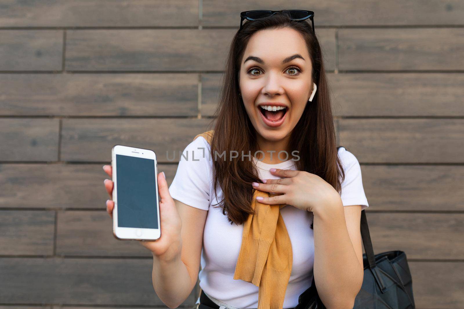 closeup attractive shocked surprised young woman wearing casual clothes standing in the street holding and showing mobile phone looking at camera. Mockup, cutout