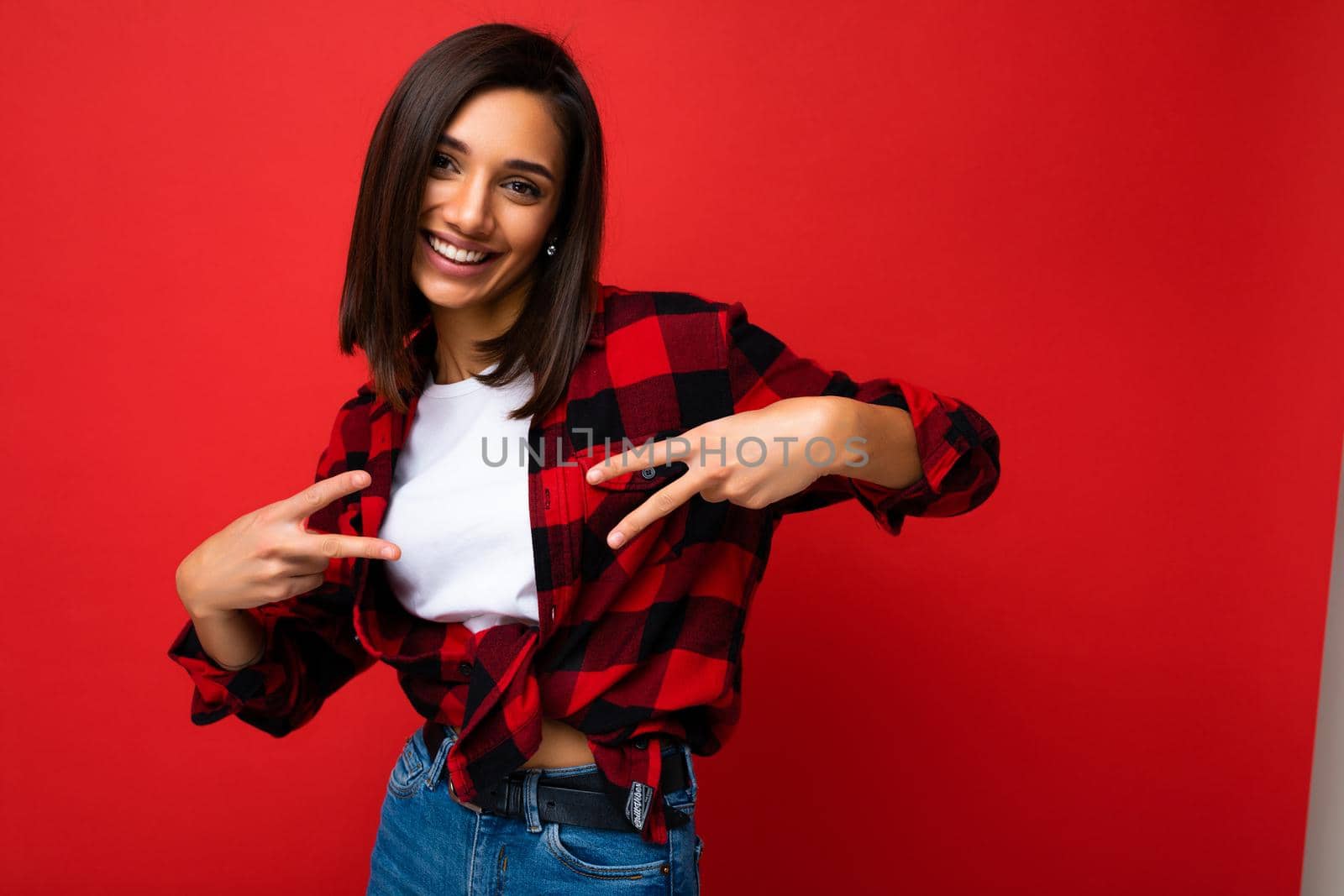 Young happy positive cool pretty brunette woman with sincere emotions wearing white t-shirt and trendy red check shirt isolated on red background with free space and having fun.