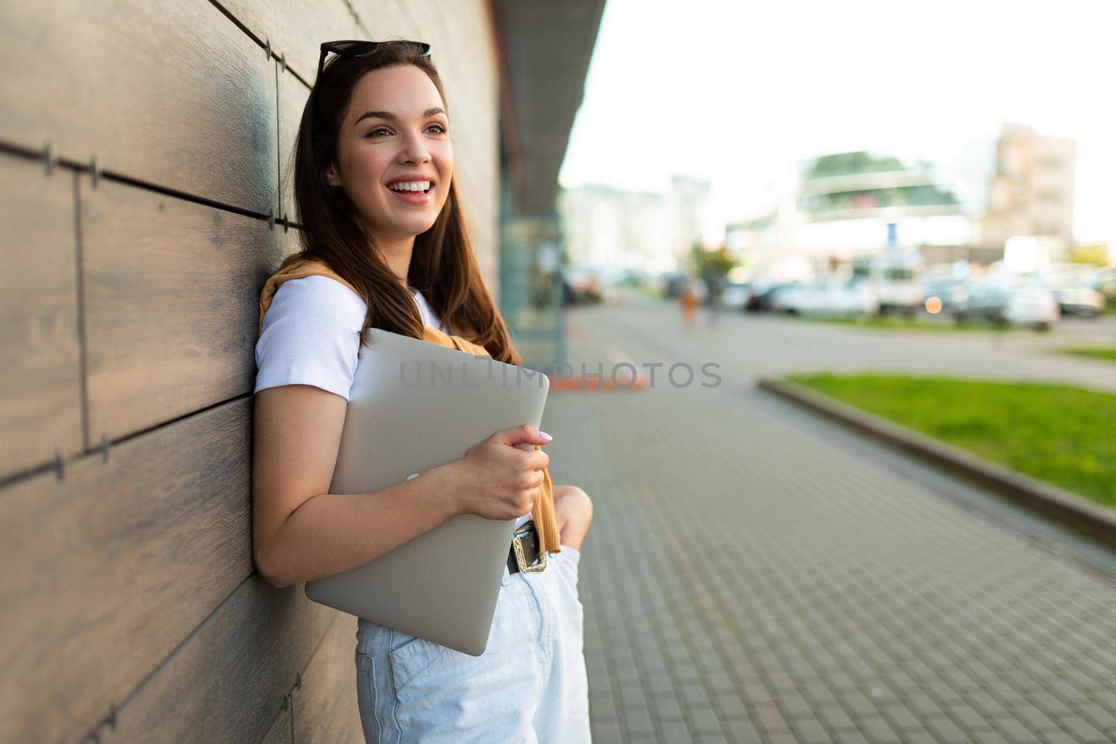 happy nice-looking charming attractive lovely fascinating cheerful cheery straight-haired brunet woman looking to side in the street with laptop computer and glasses in hands with white t-shirt and jeans.