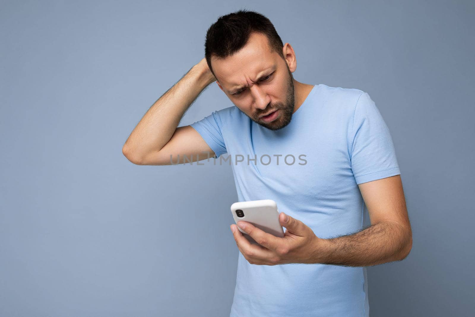 Photo shot of handsome don't understanding asking good looking young man wearing casual stylish outfit poising isolated on background with empty space holding in hand and using mobile phone messaging sms looking at smartphone display screen.