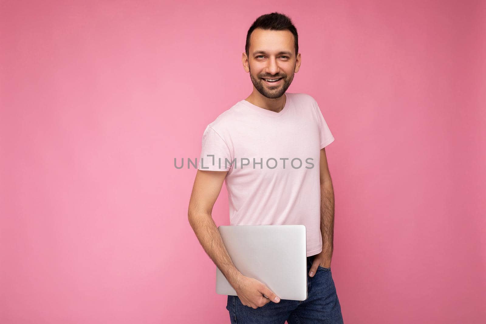 Handsome smiling brunet man holding laptop computer looking at camera in t-shirt on isolated pink background.