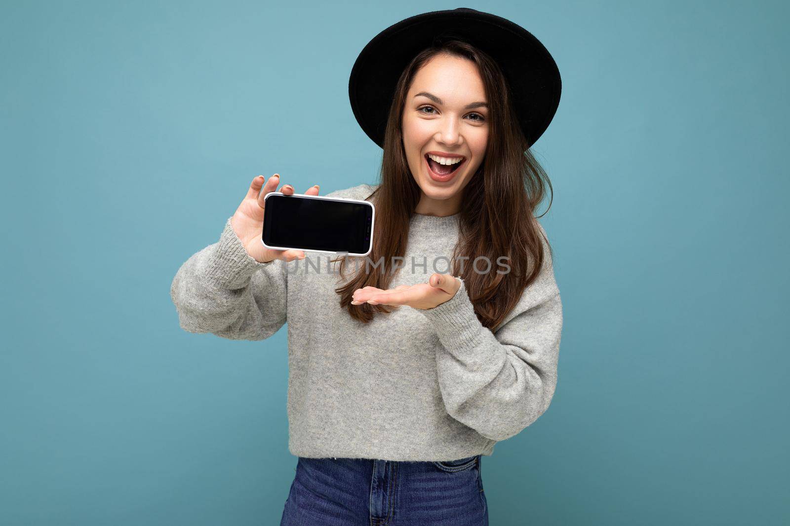 Pretty young smiling woman wearing black hat and grey sweater holding phone looking at camera isolated on background.Mock up, cutout