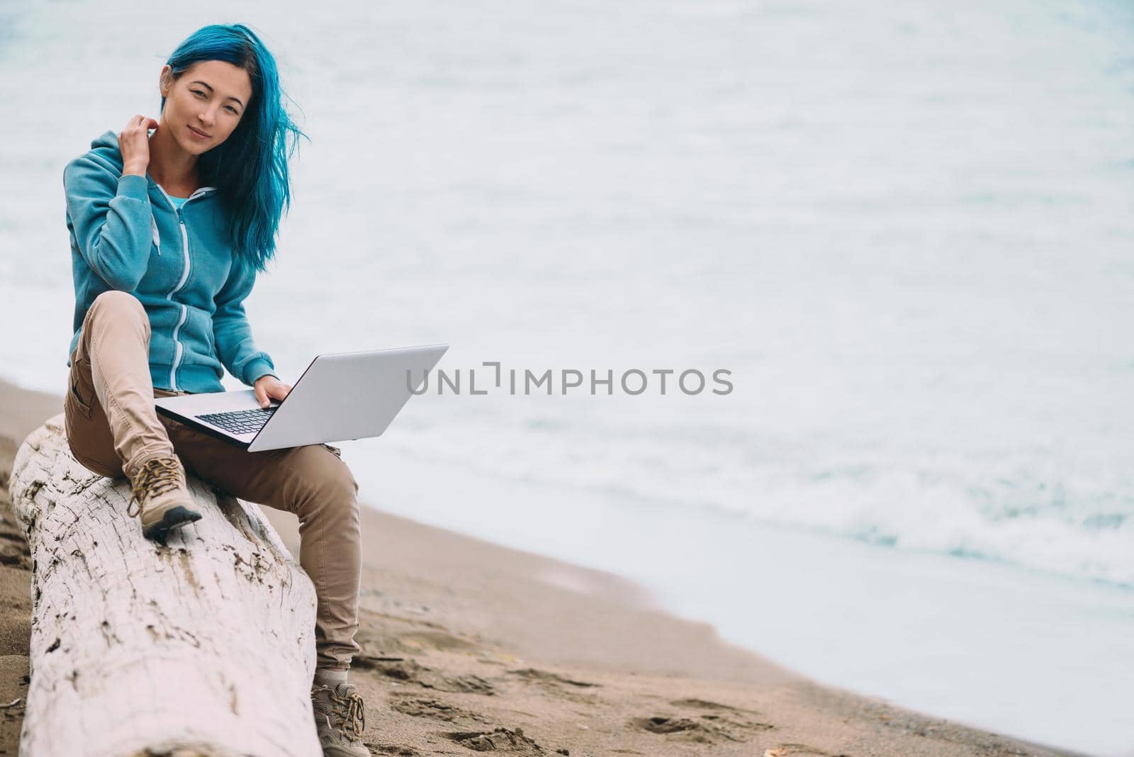Freelancer young beautiful woman sitting on beach near the sea with laptop and looking at camera. Space for text in right part of the image