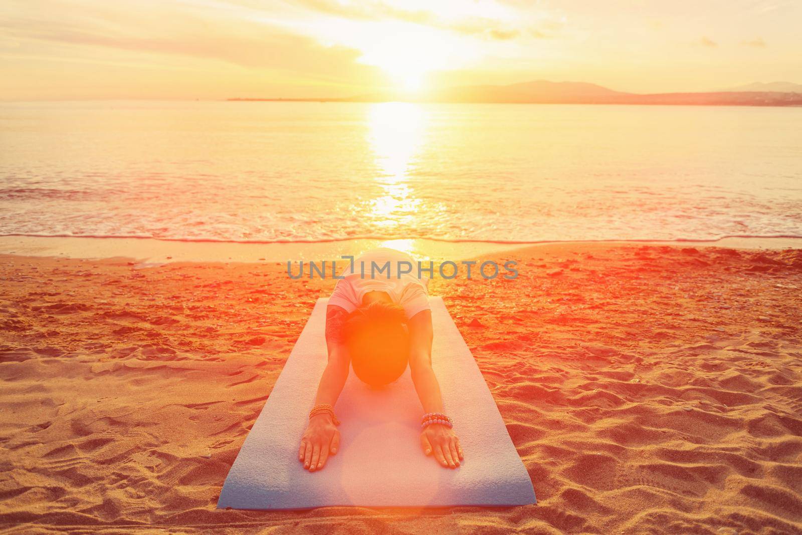 Young woman doing yoga exercise on sand beach near the sea at sunset in summer, woman stretching her back