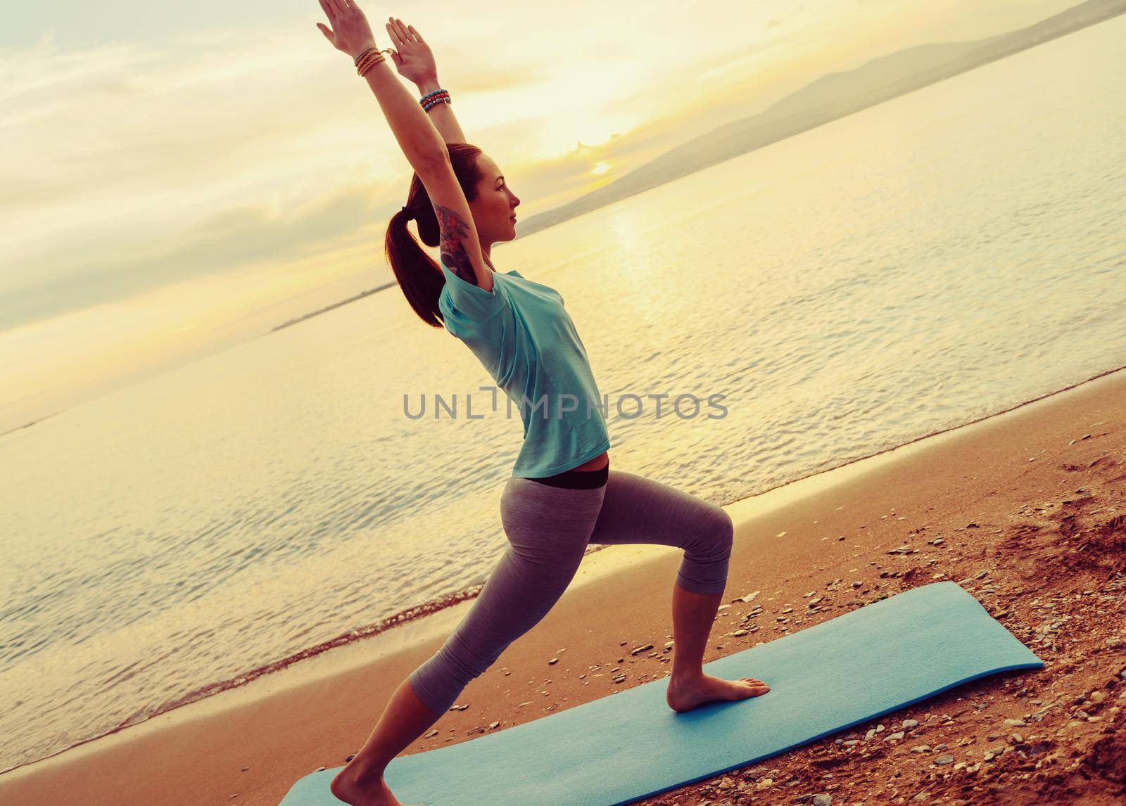 Beautiful young woman practicing yoga on mat on sand beach near the sea on sunset