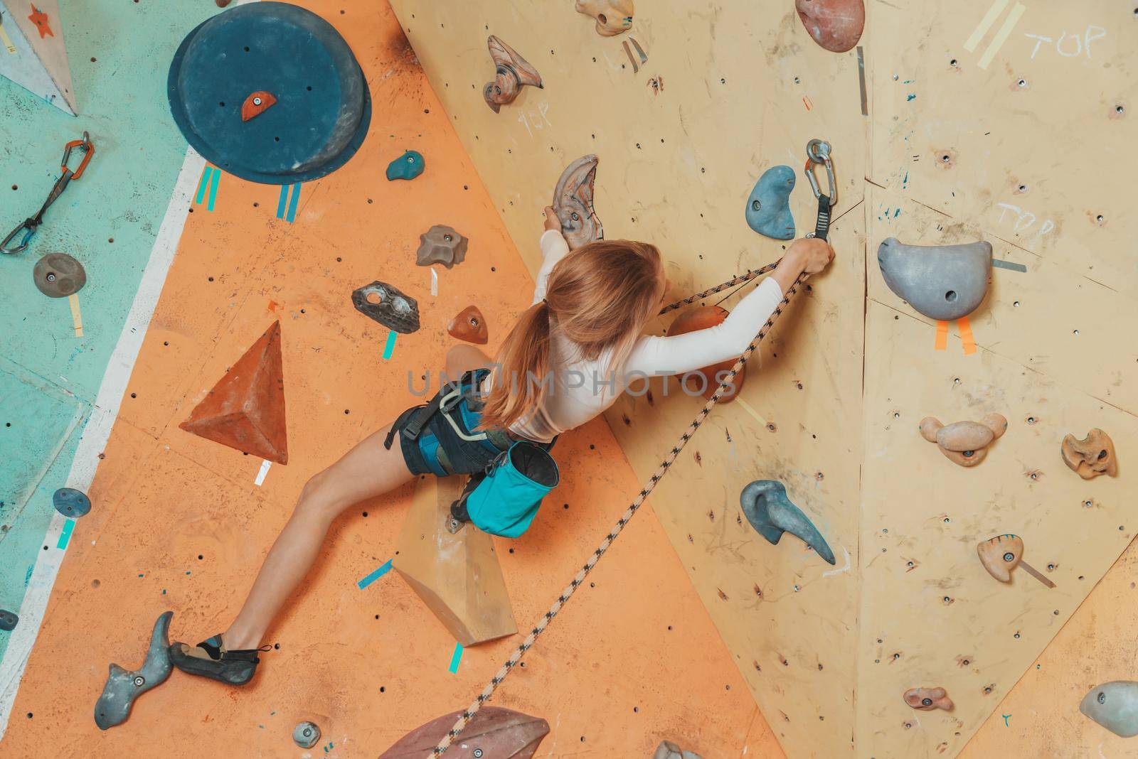 Teen girl climbing on artificial boulders of rock wall with safety harness. Girl inserting the rope in a quickdraw