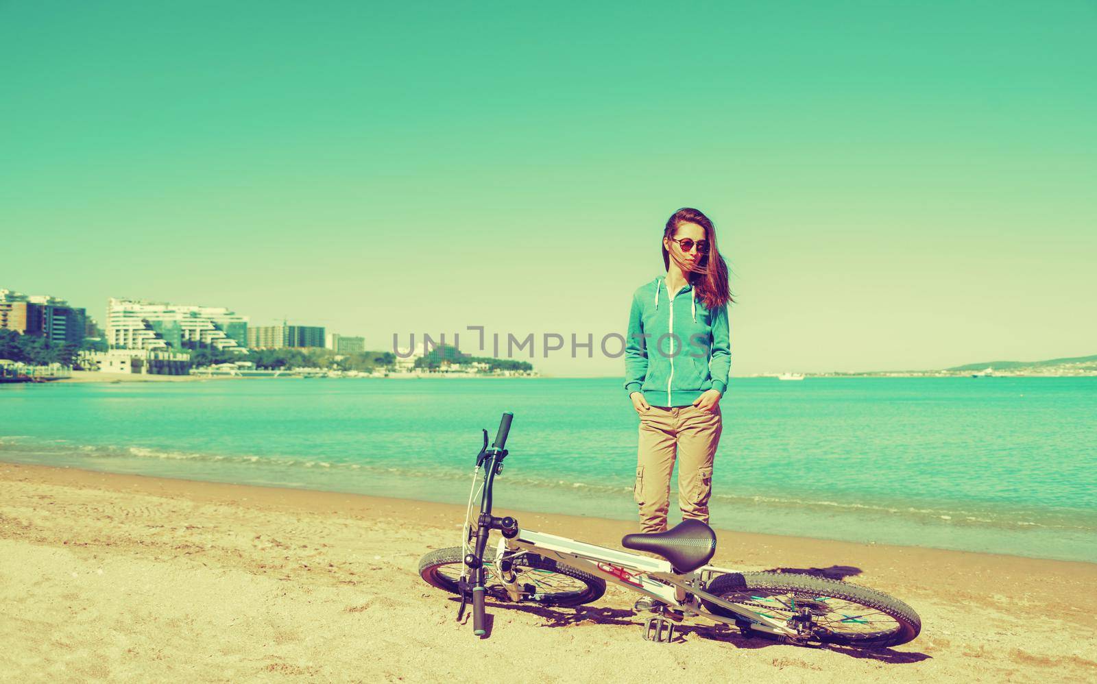 Beautiful girl standing near a bicycle on sand beach in summer in a town. Image with instagram color effect