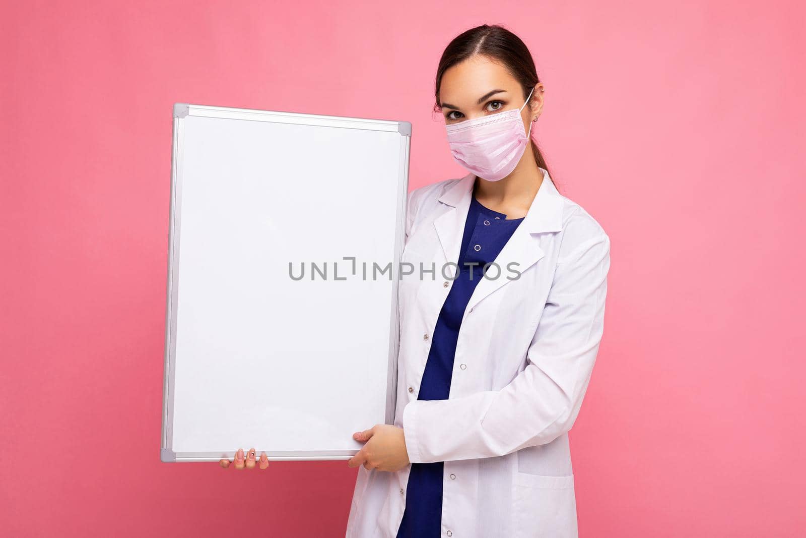 Woman doctor wearing a white medical coat and a mask holding blank board with copy space for text isolated on background. Coronavirus concept.