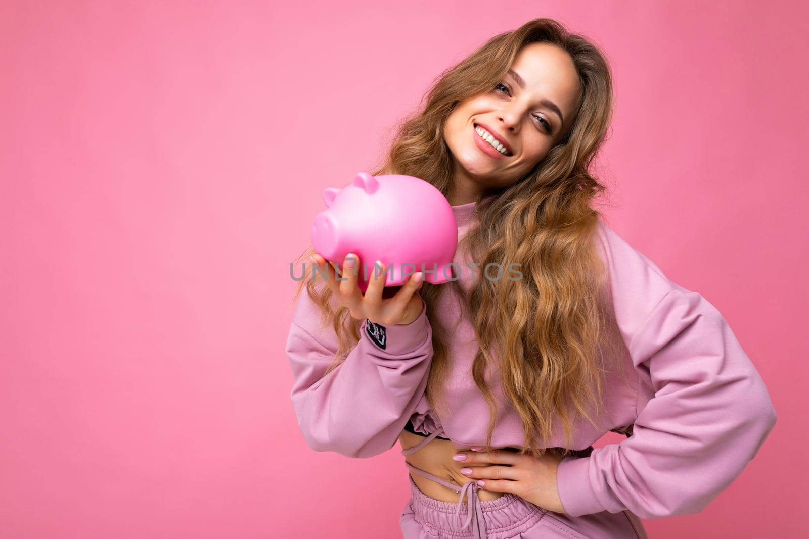 Portrait photo of happy positive smiling young beautiful attractive woman with wavy long blonde hair with sincere emotions wearing trendy pink hoodie isolated over pink background with copy space and holding pink pig penny bank. Money box concept.