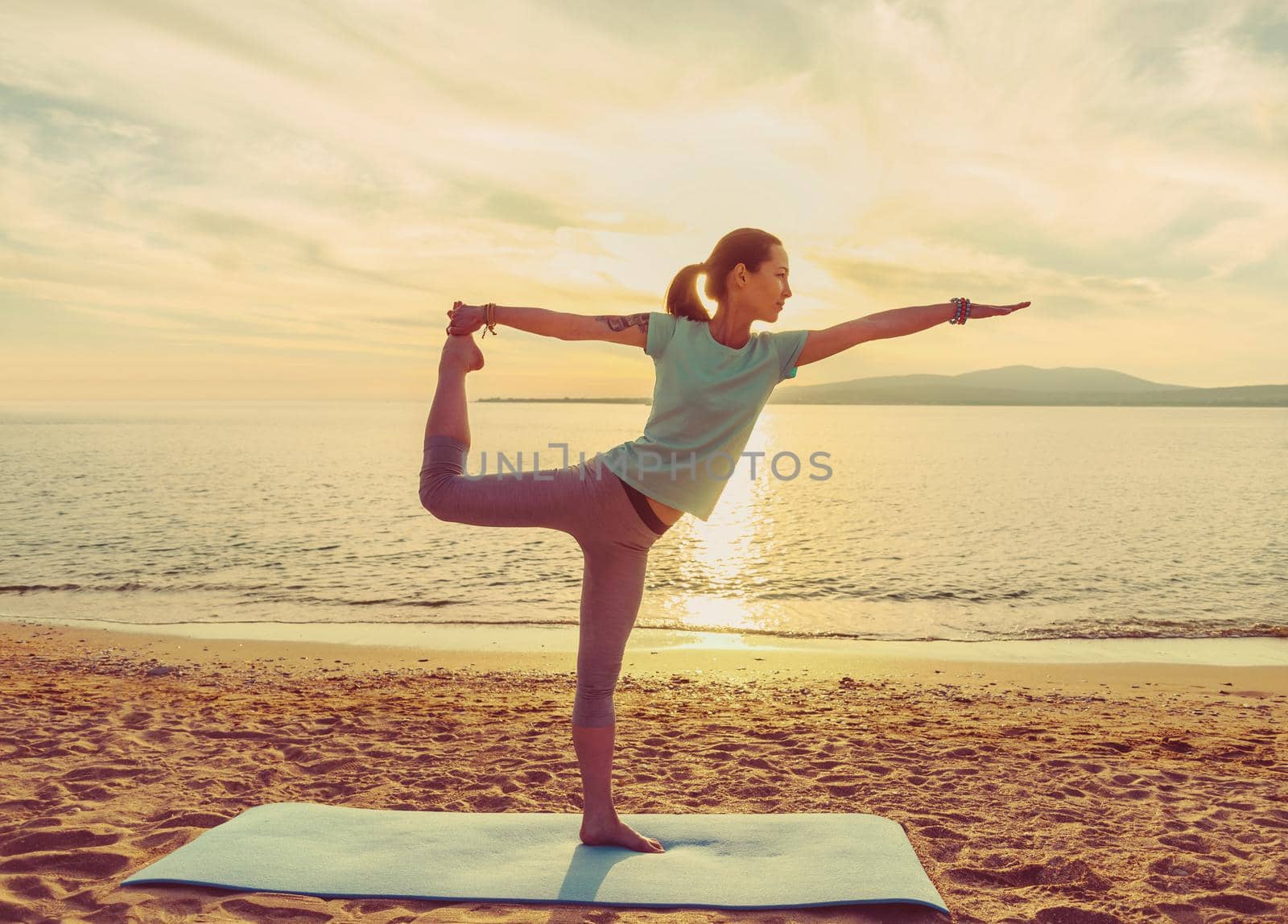 Young woman practicing yoga on mat on sand beach near the sea on sunset, woman in stretching balance pose
