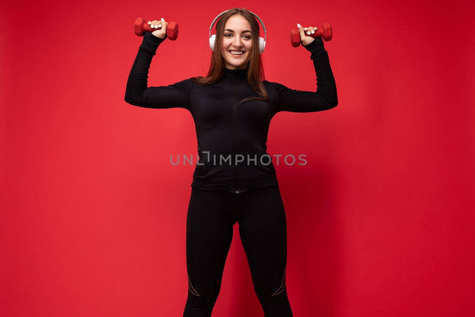 Beautiful happy smiling young brunet woman wearing black sport clothes isolated on red background doing fitness using dumbbells wearing white bluetooth headsets listening to music looking at camera.