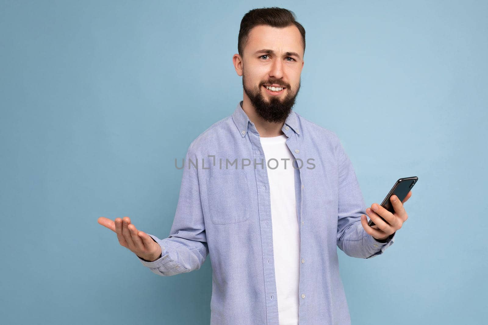 closeup Photo shot of handsome calm asking good looking young man wearing casual stylish outfit poising isolated on background with empty space holding in hand and using mobile phone messaging sms looking at smartphone display screen.